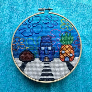 May include: Cross-stitch embroidery in a wooden hoop featuring a scene with a blue house, a pineapple, and a brown mushroom. The background is blue with bubbles. The embroidery is on a blue fabric background.