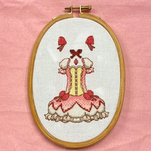 May include: A cross-stitch embroidery of a pink and yellow dress with a white background. The dress has a yellow bodice and a pink skirt with a white ruffle. The dress is framed in a wooden oval frame.