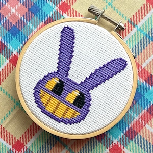 May include: A purple and yellow cross-stitch embroidery of a cartoon bunny with a wide smile, framed in a wooden hoop.