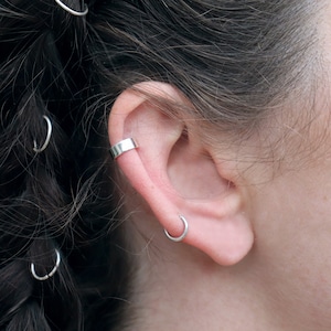 May include: Close-up of an ear with silver jewellery. A silver ear cuff is on the upper ear, and a small silver hoop earring is in the lobe. Silver hair rings are in the dark hair.