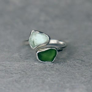 May include: A silver ring featuring two irregularly shaped, bezel-set stones. One stone is a pale green color, and the other is a vibrant green. The ring has a modern, open design, with the stones positioned on opposite sides of the band.