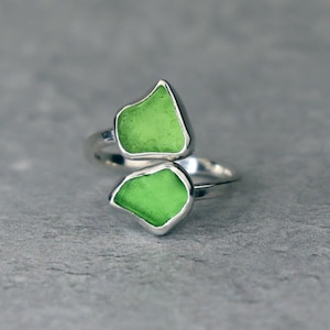 May include: A silver-toned ring featuring two irregular, bright green, sea glass-like stones. The stones are set in a bezel setting, and the ring has a wrap-around design. The ring is displayed on a gray surface.