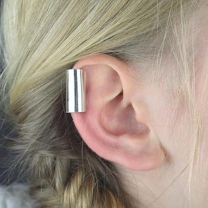 May include: A silver ear cuff is worn on the ear. The cuff is a simple, cylindrical design. The ear cuff is made of a shiny, metallic material. The ear is pink and the hair is blonde.