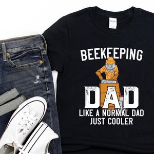 May include: Black t-shirt with the words "BEEKEEPING DAD" and an illustration of a beekeeper. The text "LIKE A NORMAL DAD JUST COOLER" is below the word "DAD". The shirt is paired with blue jeans and white sneakers.