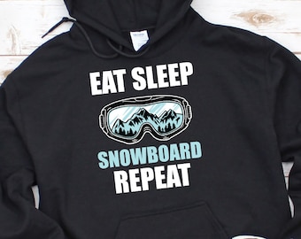 Puede incluir: Sudadera con capucha negra con el texto "Eat Sleep Snowboard Repeat" y un gráfico de unas gafas de esquí con montañas en el fondo.