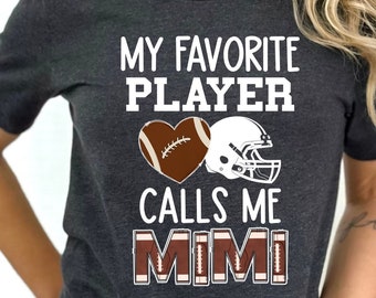 Könnte beinhalten: Dunkelgraues T-Shirt mit dem Text "MY FAVORITE PLAYER CALLS ME MIMI". Das Design beinhaltet ein herzförmiges Football und einen Footballhelm. Der Text und die Grafiken sind in Weiß und Braun.
