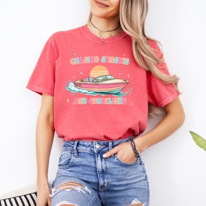 Pu&ograve; includere: T-shirt color corallo con grafica di una barca sull'acqua e un tramonto. Il testo "Chasing Sunsets + And Shorelines" &egrave; scritto in un font retr&ograve;. La maglietta &egrave; abbinata a pantaloncini di jeans blu. Tema nautico.