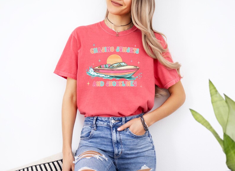 Pu&ograve; includere: T-shirt color corallo con grafica di una barca sull'acqua e un tramonto. Il testo "Chasing Sunsets + And Shorelines" &egrave; scritto in un font retr&ograve;. La maglietta &egrave; abbinata a pantaloncini di jeans blu. Tema nautico.