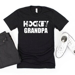 May include: Black t-shirt with white text that reads "HOCKEY GRANDPA" with hockey player silhouettes in the letters.
