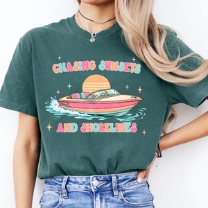 Pu&ograve; includere: T-shirt verde scuro con una grafica colorata di una barca sull'acqua, un sole al tramonto e la scritta "Chasing Sunsets and Shorelines". Il design include stelle e colori pastello.
