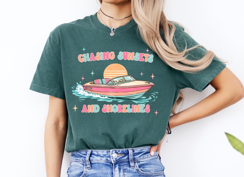 Pu&ograve; includere: T-shirt verde scuro con una grafica colorata di una barca sull'acqua, un sole al tramonto e la scritta "Chasing Sunsets and Shorelines". Il design include stelle e colori pastello.