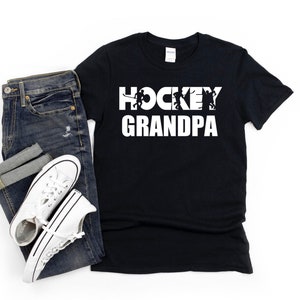 May include: Black t-shirt with white text that reads "HOCKEY GRANDPA" with a silhouette of hockey players in the word "HOCKEY".