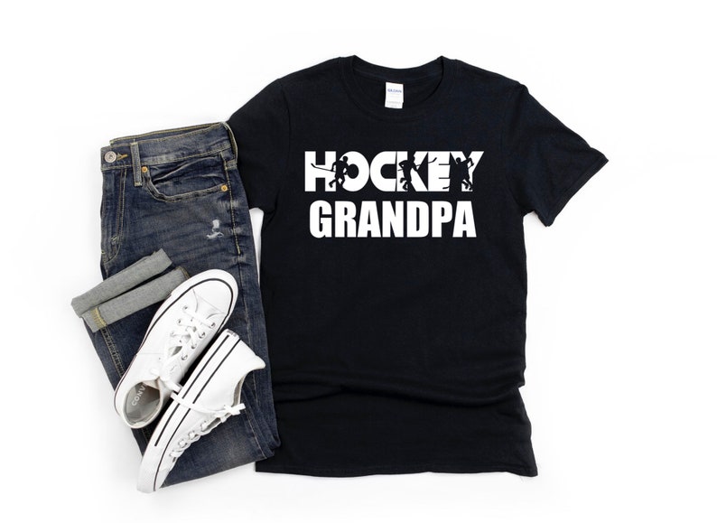 May include: Black t-shirt with white text that reads "HOCKEY GRANDPA" with a silhouette of hockey players in the word "HOCKEY".