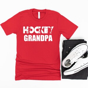 May include: Red t-shirt with white text that says "Hockey Grandpa" with a graphic of hockey players in the word "Hockey".