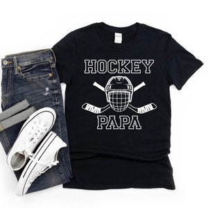 May include: Black t-shirt with the words "HOCKEY PAPA" in white block letters, along with a hockey helmet and crossed hockey sticks. The shirt is paired with blue jeans and white sneakers.