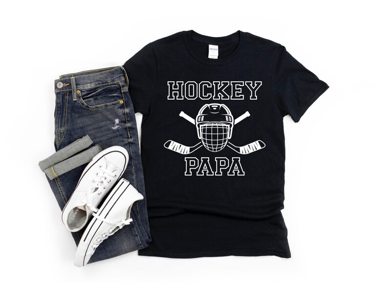 May include: Black t-shirt with the words "HOCKEY PAPA" in white block letters, along with a hockey helmet and crossed hockey sticks. The shirt is paired with blue jeans and white sneakers.