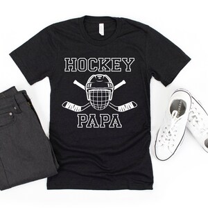 May include: Black t-shirt with the words "HOCKEY PAPA" in white block letters, along with a hockey helmet and crossed hockey sticks. The shirt is paired with dark jeans and white sneakers.