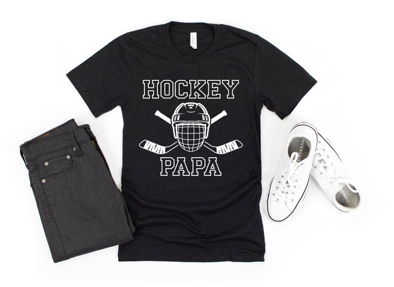 May include: Black t-shirt with the words "HOCKEY PAPA" in white block letters, along with a hockey helmet and crossed hockey sticks. The shirt is paired with dark jeans and white sneakers.