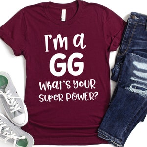 May include: A maroon t-shirt with white text that reads "I'm a GG What's your super power?" The shirt is laid flat on a white surface with a pair of gray sneakers and a pair of blue jeans.