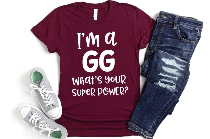 May include: A maroon t-shirt with white text that reads "I'm a GG What's your super power?" The shirt is laid flat on a white surface with a pair of gray sneakers and a pair of blue jeans.
