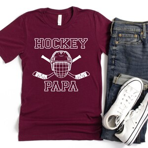 May include: A maroon t-shirt with the words "HOCKEY PAPA" in white block letters, along with a hockey helmet and crossed hockey sticks. The shirt is paired with blue jeans and white sneakers.