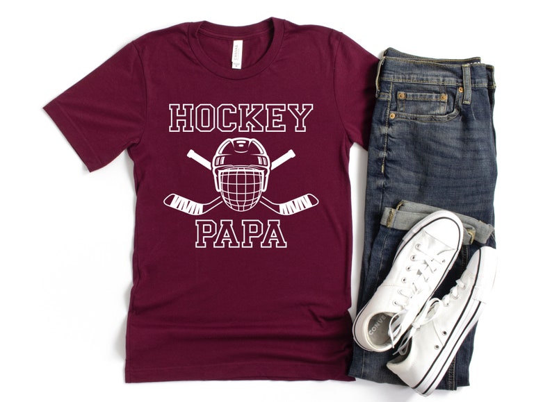 May include: A maroon t-shirt with the words "HOCKEY PAPA" in white block letters, along with a hockey helmet and crossed hockey sticks. The shirt is paired with blue jeans and white sneakers.