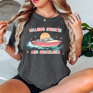 Pu&ograve; includere: T-shirt grigio scuro con la scritta "Chasing Sunsets and Shorelines" sopra una grafica di una barca rosa e gialla. Il design include un tramonto e delle stelle. Un capo d'abbigliamento casual.