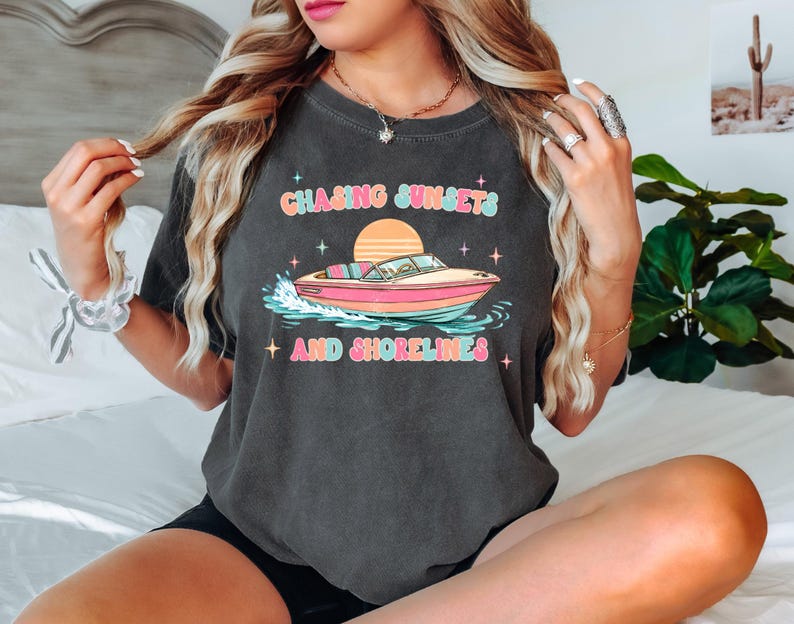 Pu&ograve; includere: T-shirt grigio scuro con la scritta "Chasing Sunsets and Shorelines" sopra una grafica di una barca rosa e gialla. Il design include un tramonto e delle stelle. Un capo d'abbigliamento casual.