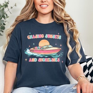 Pu&ograve; includere: T-shirt blu navy con una grafica colorata di una barca sull'acqua, un tramonto e la scritta "Chasing Sunsets and Shorelines". Il design presenta colori pastello e stelle. &Egrave; un capo di abbigliamento casual.