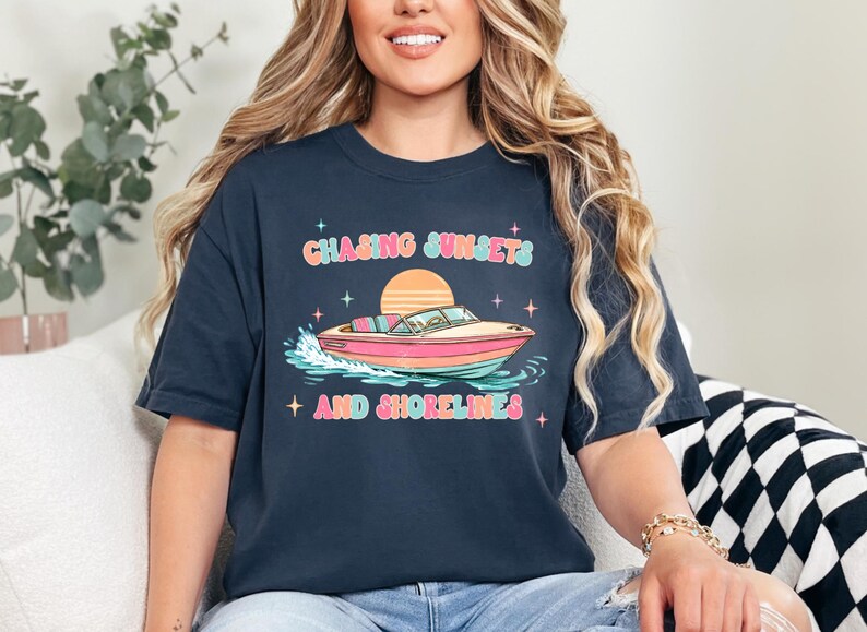 Pu&ograve; includere: T-shirt blu navy con una grafica colorata di una barca sull'acqua, un tramonto e la scritta "Chasing Sunsets and Shorelines". Il design presenta colori pastello e stelle. &Egrave; un capo di abbigliamento casual.