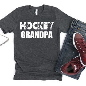 May include: A dark grey t-shirt with the text "HOCKEY GRANDPA" in white. The text is overlaid with a silhouette of hockey players.