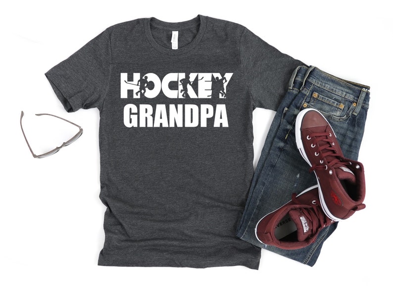 May include: A dark grey t-shirt with the text "HOCKEY GRANDPA" in white. The text is overlaid with a silhouette of hockey players.