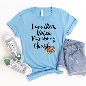 K&ouml;nnte beinhalten: Ein hellblaues T-Shirt mit dem Text "Ich bin ihre Stimme Sie sind mein Herz" und einem Herz aus Puzzle-Teilen in den Farben des Autismus-Bandes.