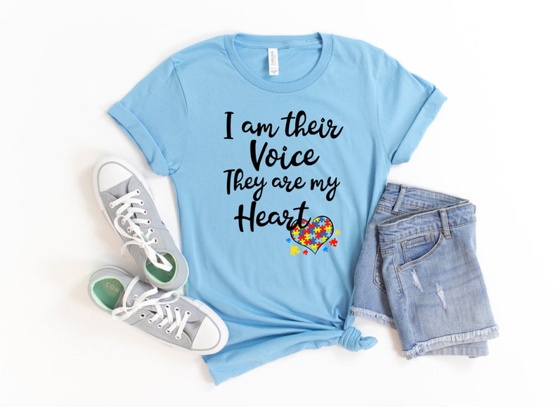K&ouml;nnte beinhalten: Ein hellblaues T-Shirt mit dem Text "Ich bin ihre Stimme Sie sind mein Herz" und einem Herz aus Puzzle-Teilen in den Farben des Autismus-Bandes.
