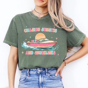 Pu&ograve; includere: T-shirt verde oliva con grafica colorata di una barca sull'acqua, un tramonto e la scritta "Chasing Sunsets and Shorelines". Il design presenta colori pastello e accenti di stelle.