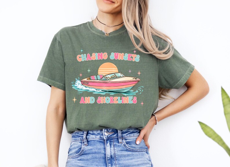 Pu&ograve; includere: T-shirt verde oliva con grafica colorata di una barca sull'acqua, un tramonto e la scritta "Chasing Sunsets and Shorelines". Il design presenta colori pastello e accenti di stelle.
