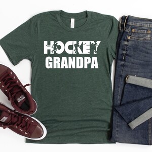 May include: A dark green t-shirt with the text "HOCKEY GRANDPA" in white. The text is surrounded by silhouettes of hockey players. The t-shirt is laid flat on a white surface with a pair of brown high-top sneakers and a pair of blue jeans.