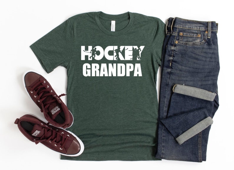 May include: A dark green t-shirt with the text "HOCKEY GRANDPA" in white. The text is surrounded by silhouettes of hockey players. The t-shirt is laid flat on a white surface with a pair of brown high-top sneakers and a pair of blue jeans.