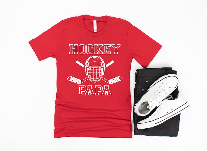 May include: Red t-shirt with the words "HOCKEY PAPA" in white block letters, along with a hockey helmet and crossed hockey sticks. The shirt is paired with black jeans and white sneakers.