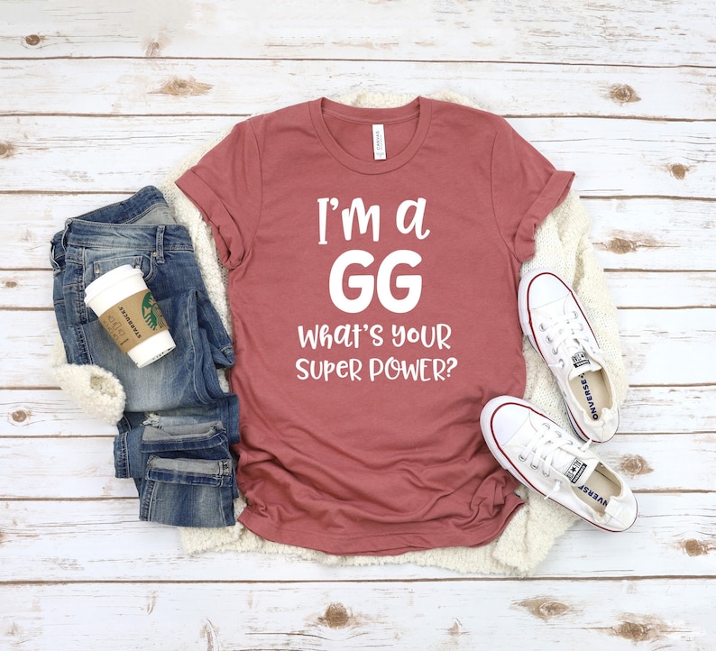 May include: A maroon t-shirt with the text "I'm a GG What's your super power?" in white. The shirt is laid out on a wooden surface with a pair of white sneakers and a pair of blue jeans.