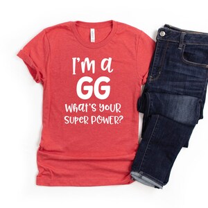 May include: Red t-shirt with white text that reads "I'm a GG What's your super power?" The shirt is folded and laid on a white surface next to a pair of blue jeans.