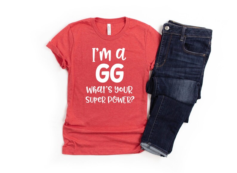 May include: Red t-shirt with white text that reads "I'm a GG What's your super power?" The shirt is folded and laid on a white surface next to a pair of blue jeans.