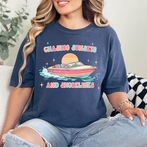 Pu&ograve; includere: T-shirt blu navy con la scritta "Chasing Sunsets and Shorelines" sopra una grafica di una barca rosa e bianca su acqua blu e verde. La maglietta ha una vestibilit&agrave; ampia.