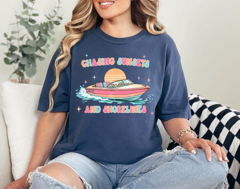 Pu&ograve; includere: T-shirt blu navy con la scritta "Chasing Sunsets and Shorelines" sopra una grafica di una barca rosa e bianca su acqua blu e verde. La maglietta ha una vestibilit&agrave; ampia.