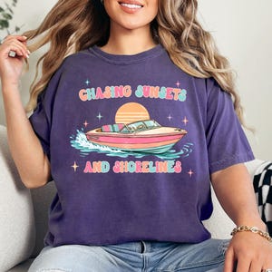 Pu&ograve; includere: Maglietta viola con grafica di una barca sull'acqua, un tramonto e la scritta "Chasing Sunsets and Shorelines" in un font retr&ograve;. La barca &egrave; rosa, arancione e blu.