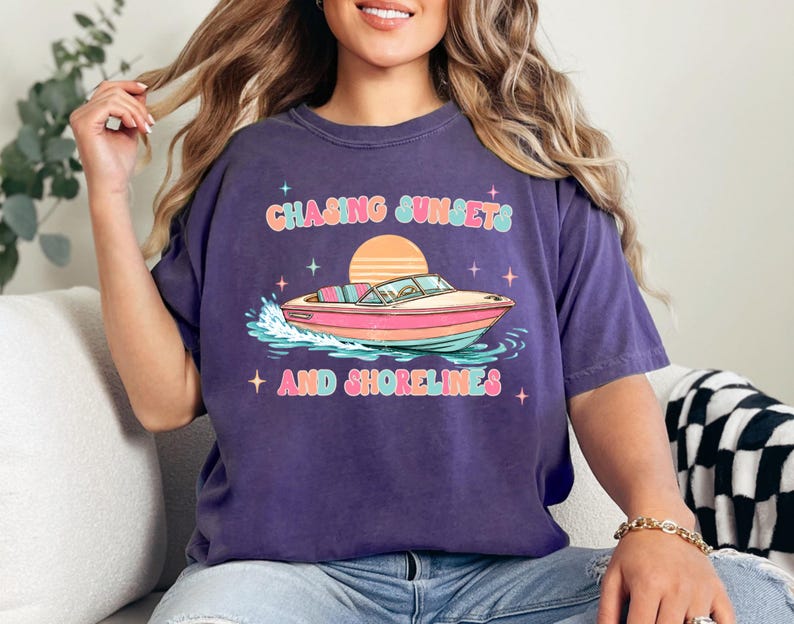 Pu&ograve; includere: Maglietta viola con grafica di una barca sull'acqua, un tramonto e la scritta "Chasing Sunsets and Shorelines" in un font retr&ograve;. La barca &egrave; rosa, arancione e blu.