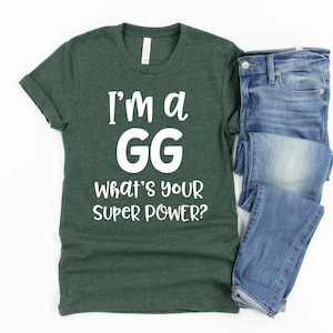 May include: A dark green t-shirt with white text that reads "I'm a GG What's your super power?" The t-shirt is folded and laid on a white surface next to a pair of blue jeans.