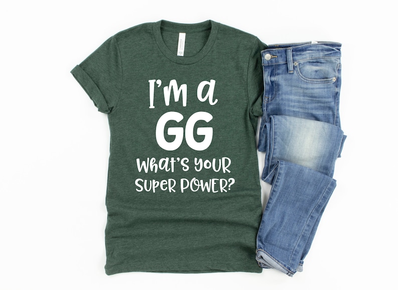 May include: A dark green t-shirt with white text that reads "I'm a GG What's your super power?" The t-shirt is folded and laid on a white surface next to a pair of blue jeans.
