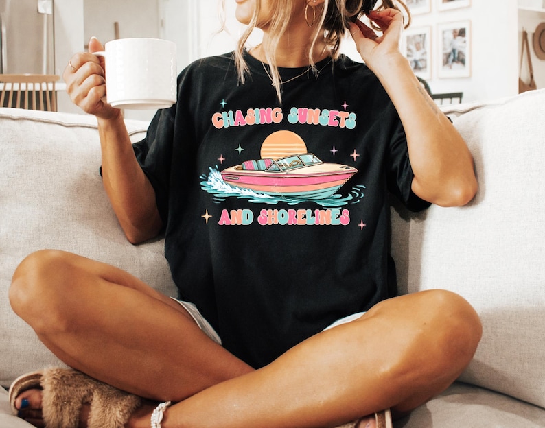 Pu&ograve; includere: T-shirt nero con una grafica colorata di una barca sull'acqua, un sole al tramonto e la scritta "Chasing Sunsets and Shorelines". Il design presenta tonalit&agrave; rosa, arancioni, blu e verdi. La maglietta &egrave; indossata da una persona.