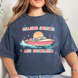 Pu&ograve; includere: T-shirt blu navy con grafica colorata di una barca sull'acqua, un tramonto e la scritta "Chasing Sunsets and Shorelines". Il design utilizza colori pastello e stelle.